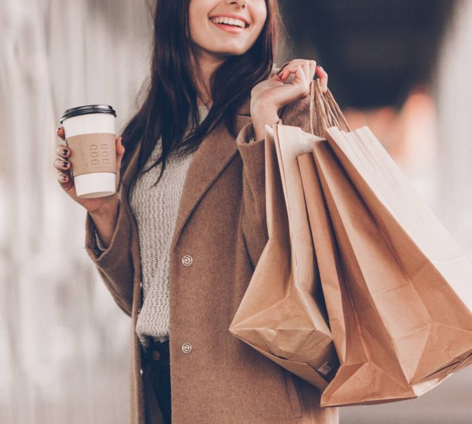 Allers Landing Smiling woman holding coffee cup and carrying several paper shopping bags while walking outdoors.