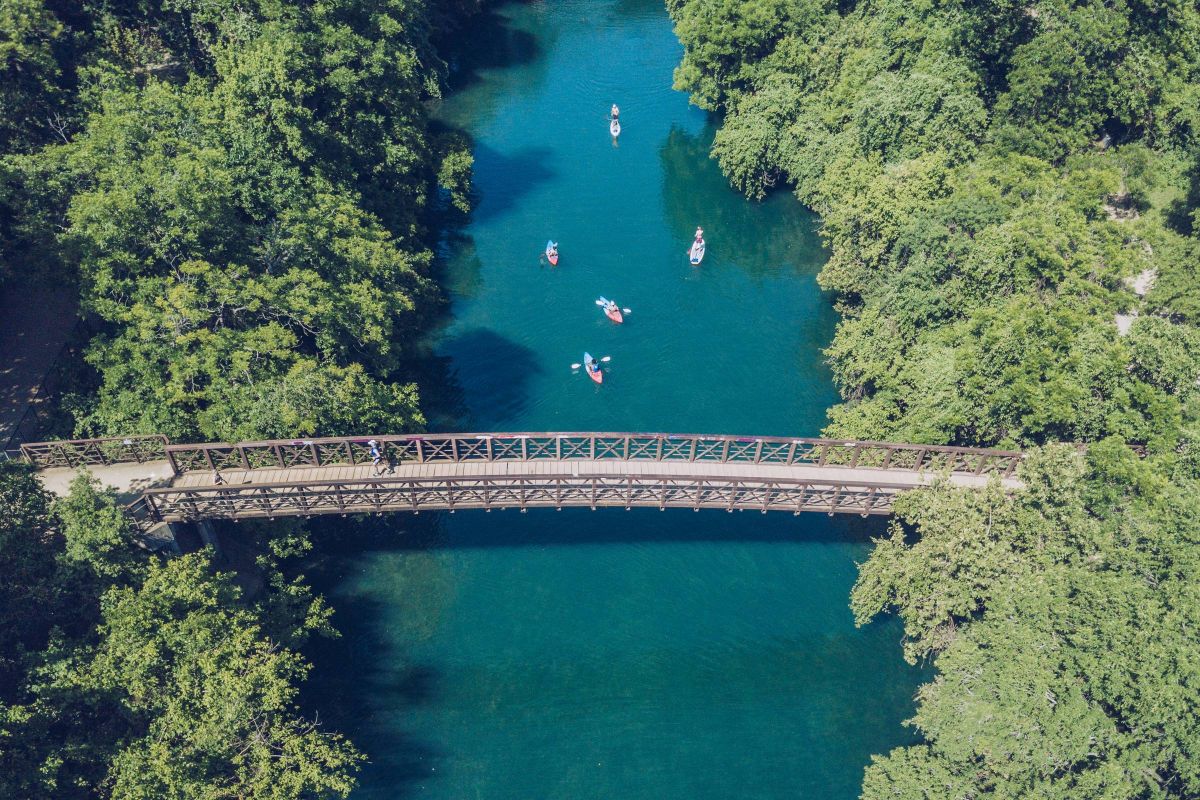 Allers Landing Aerial view of kayakers paddling under a wooden bridge on a river surrounded by lush green trees.