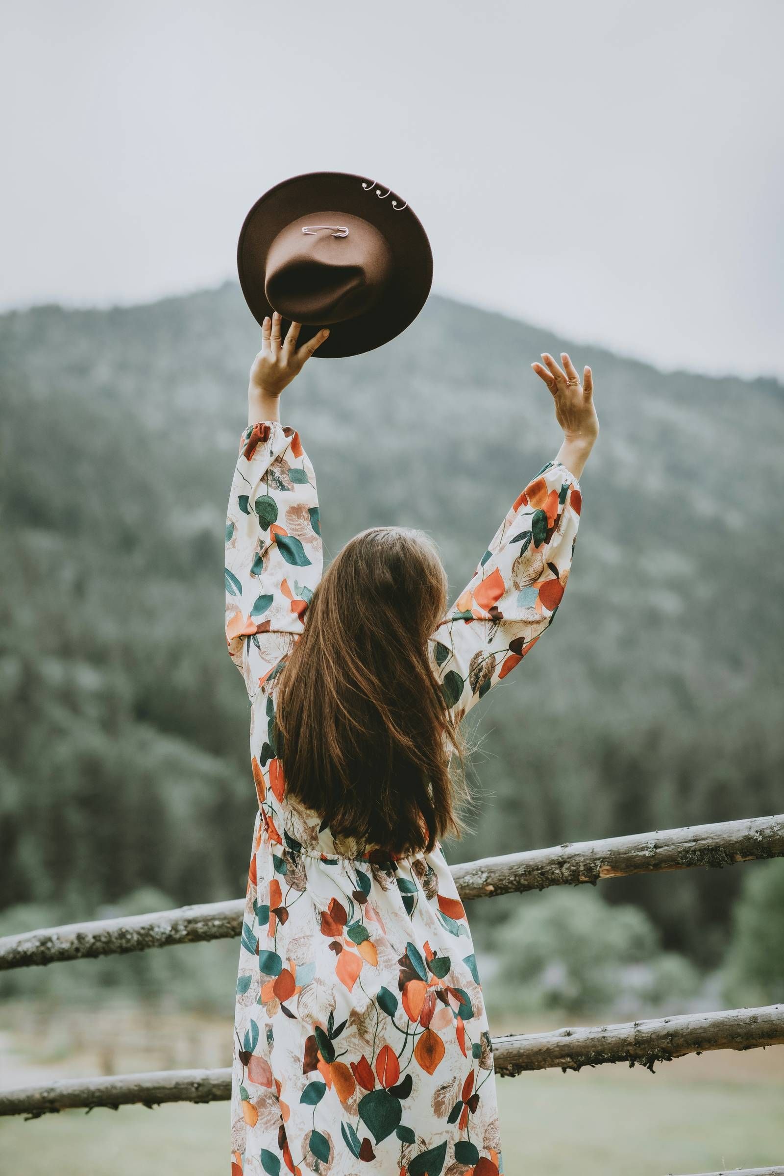 Allers Landing Woman in a floral dress stands by a fence, raising a hat toward a forested mountain in the background.