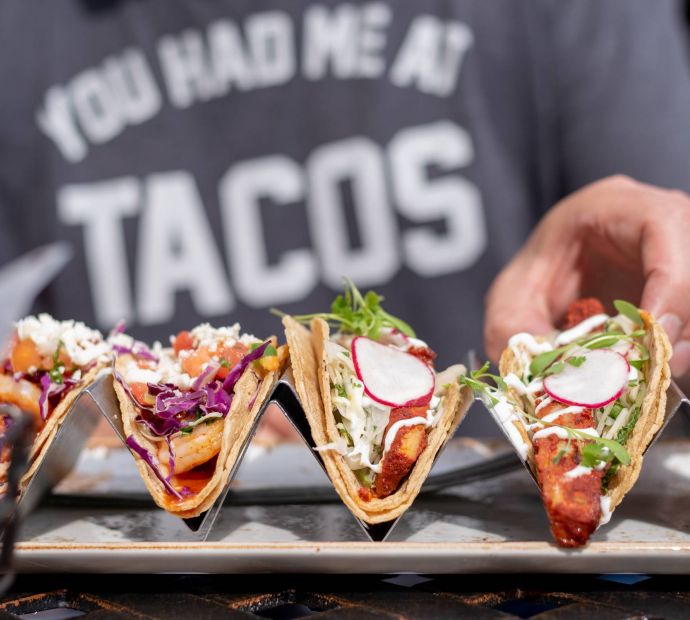 Allers Landing A person grabs a taco from a tray with four tacos; their shirt reads "You had me at TACOS.